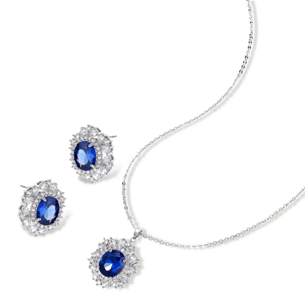 White gold plated jewelry set with blue cubic zirconia centers surrounded by clear crystal halos on cream background.
