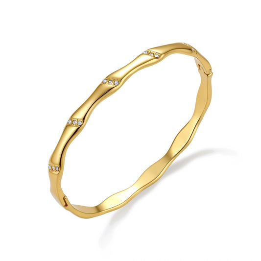 Gold-toned wavy bangle with bamboo-style segments and CZ crystal clusters on cream background. 18K plated stainless steel.